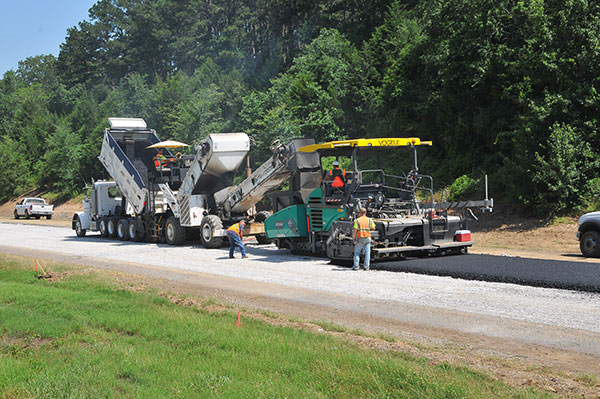 An asphalt paving train makes its way along the pulverized concrete base. Multiple layers of asphalt are required to build up the roadway and support years of constant traffic.